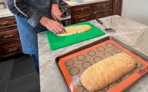 Hands cutting biscotti into slices in a kitchen. A whole biscotti loaf is in the foreground