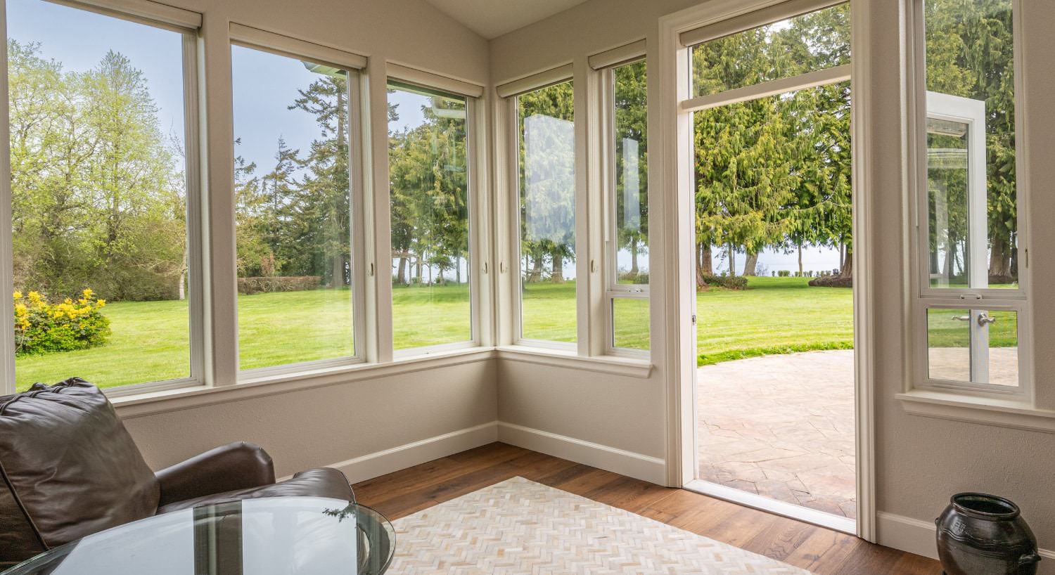 Room with light colored walls, hardwood flooring, dark leather chair, glass-top table, and multiple windows with views of the backyard