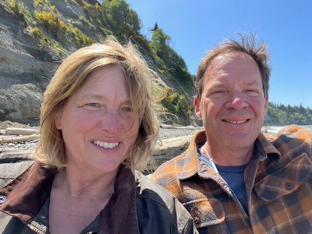 a smiling woman and man in front a cliff along the water.