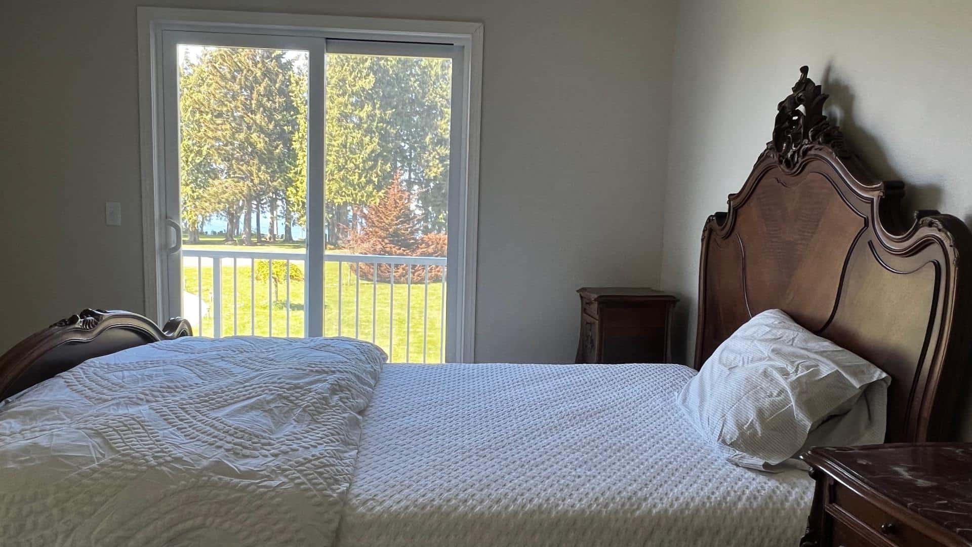 Bedroom with white walls, antique dark wooden headboard, white bedding, and sliding door to backyard