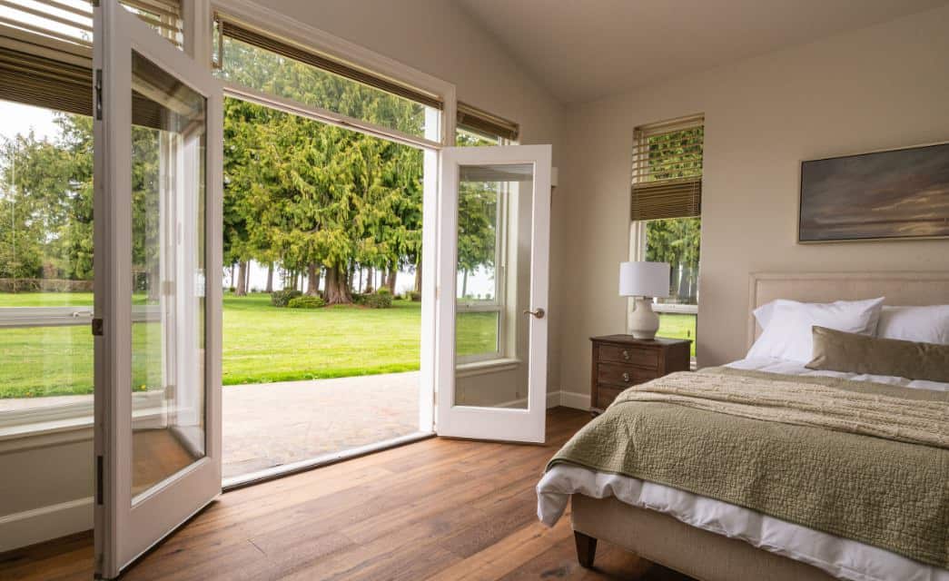 Bedroom with light colored walls, hardwood flooring, light upholstered bed with white and sage green bedding, and opened french doors to the large backyard