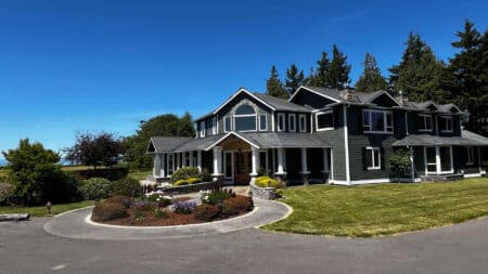 A sprawling green house with white trim and columns, surrounded by green grass, bushes and pine trees, with a round flower bed in the driveway.