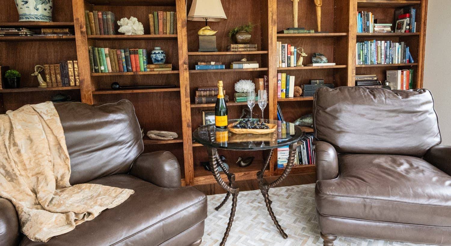 Library with light colored walls, hardwood flooring, wooden shelves filled with books, two leather armchairs, and glass-top table