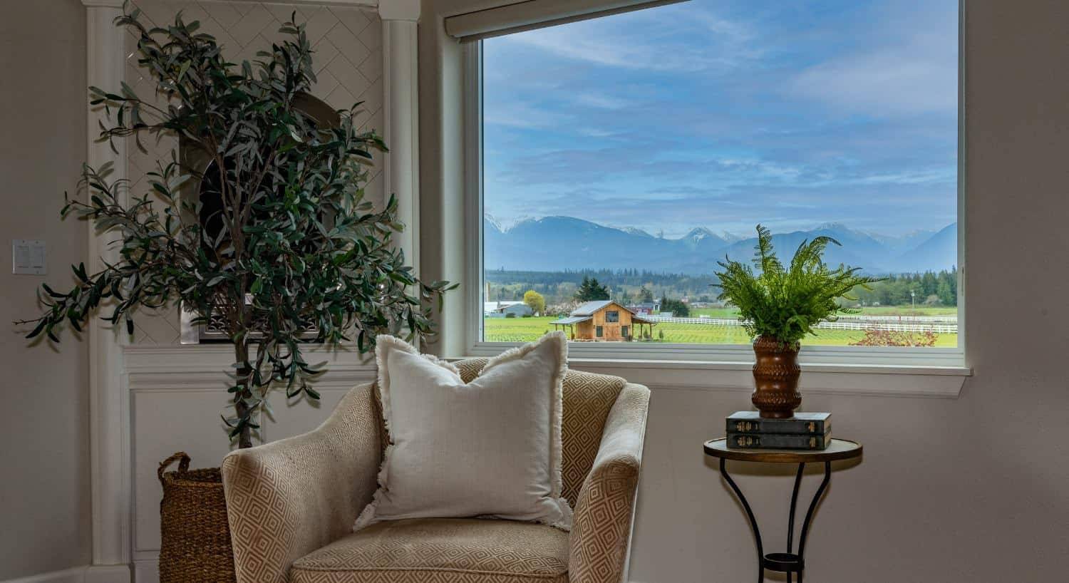 Sitting area with light colored upholstered chair, small wooden and wrought iron table, and view of the mountains from the window