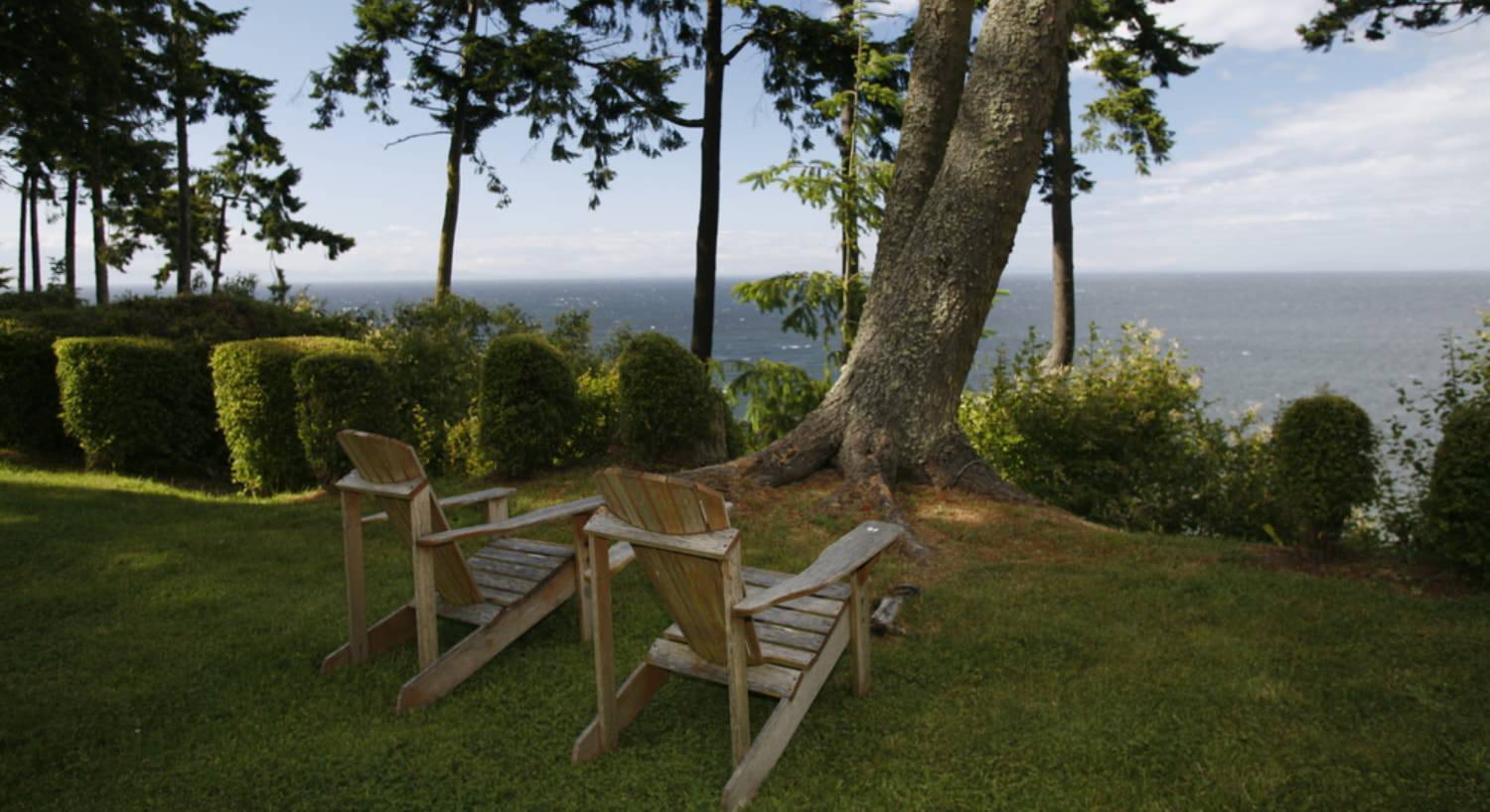 Two Adirondack chairs on green grass next to green bushes and trees looking out over the water