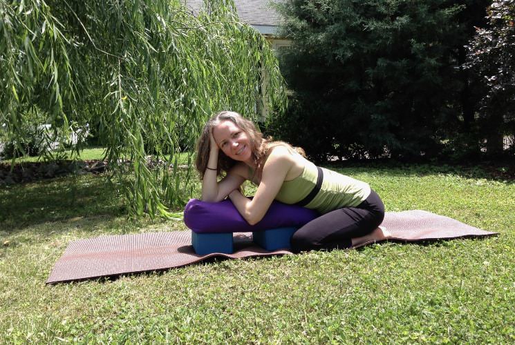 Woman in yoga attire leaning on equipment sitting on a yoga mat laying in green grass